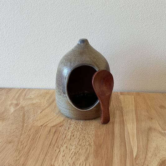 This image features a charming ceramic salt jar, crafted with a rustic, wood-fired appearance. The jar is rounded and has a wide opening, complemented by a simple wooden spoon resting against it.