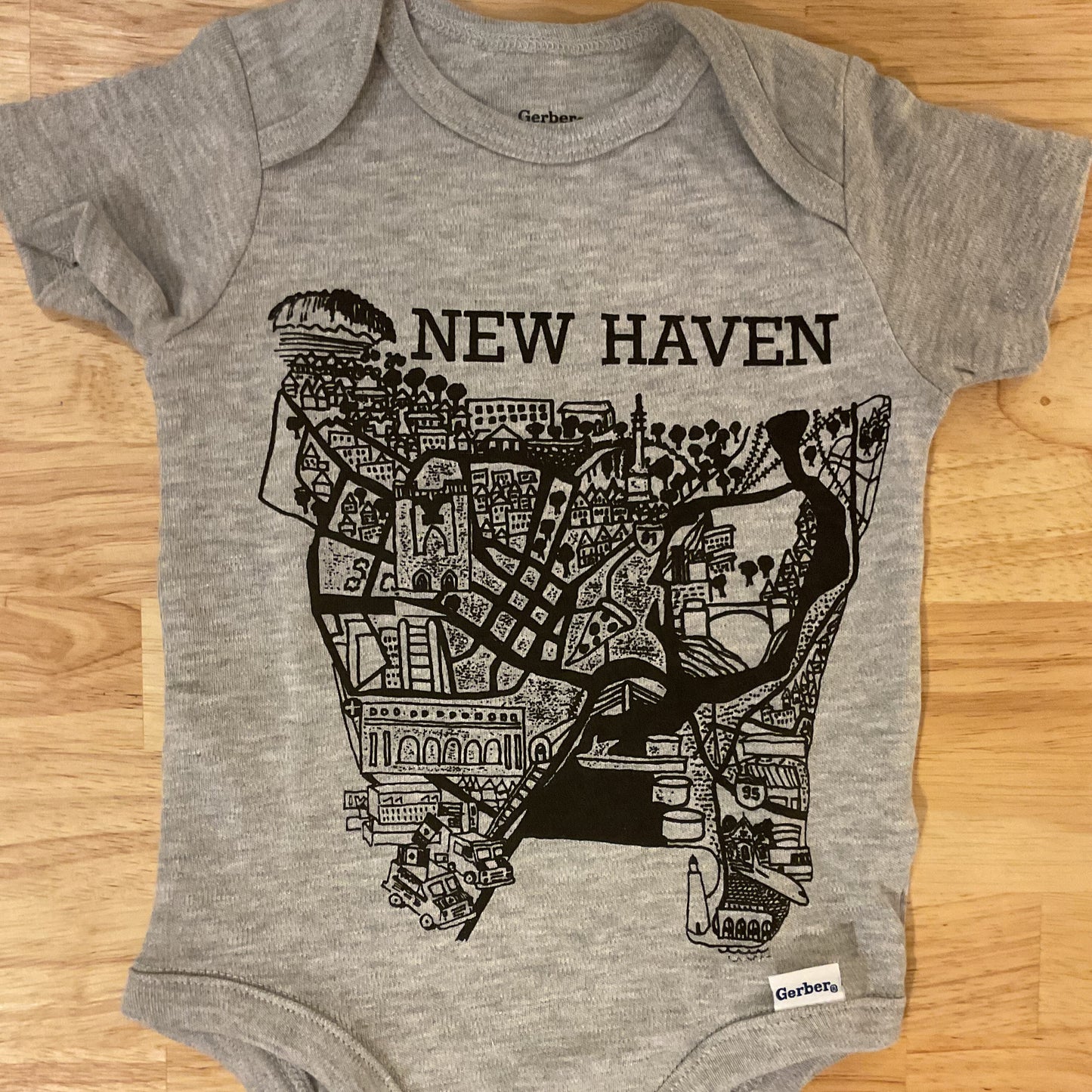 Gray baby onesie with black map design and 'New Haven' text on a wooden surface
