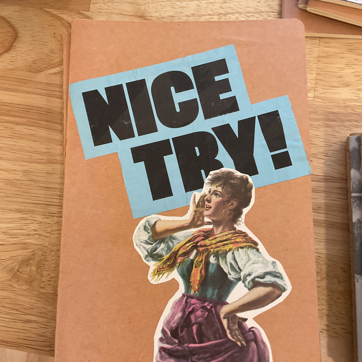 A square kraft paper card or notebook cover featuring a retro-style illustration of a woman in a purple dress and a light blue sticker that reads 'NICE TRY!' in bold black letters, placed on a wooden tabletop.