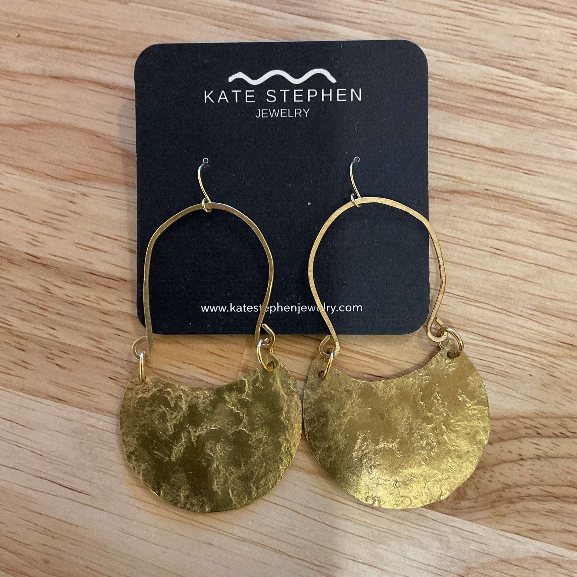 Gold earrings on a Katie Stephen Jewelry card on a wooden surface