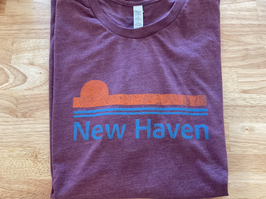 A folded heathered purple t-shirt with a retro sunset graphic in orange and blue, featuring the text 'New Haven' printed below, laid flat on a light wood floor.