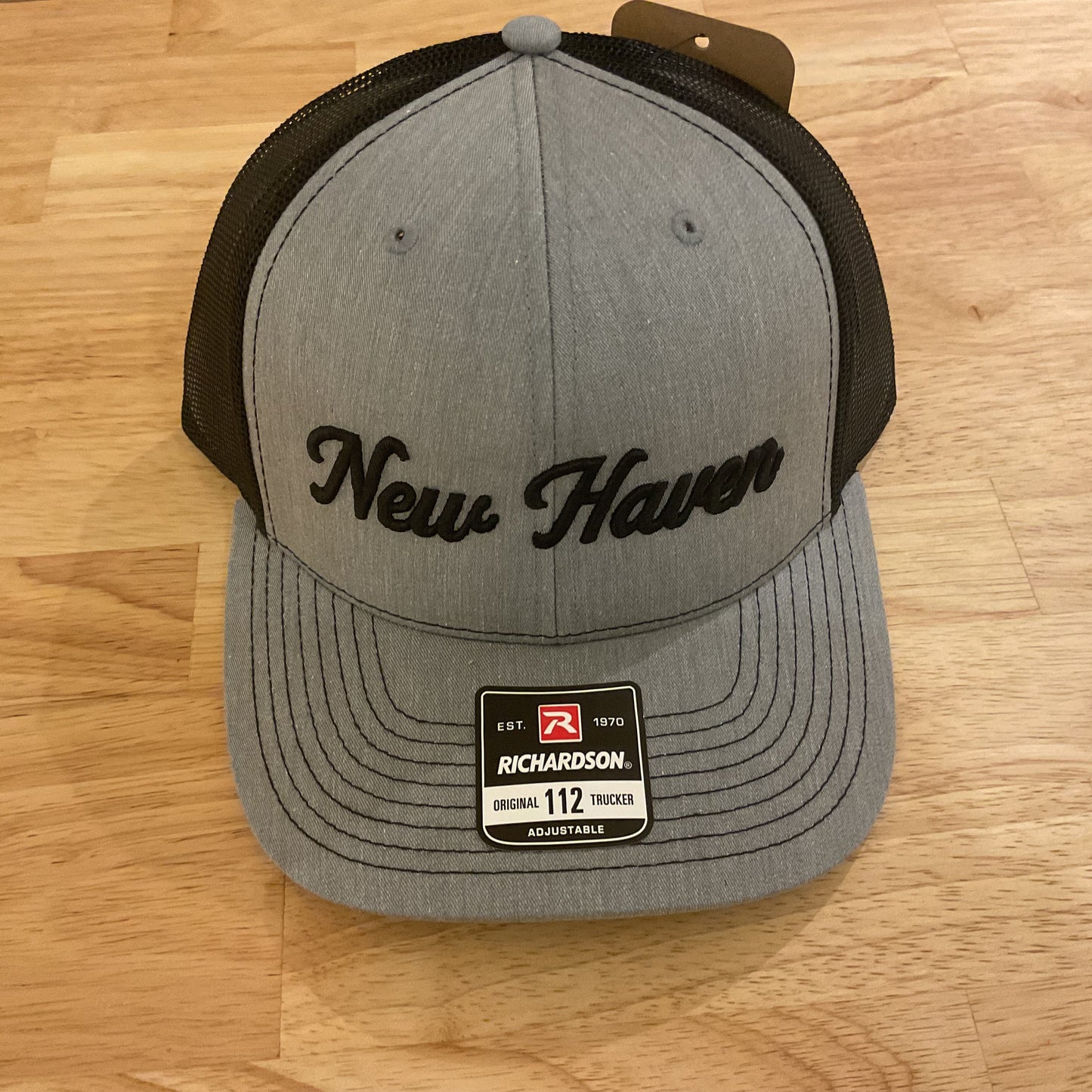 A heather gray and black Richardson 112 trucker hat with 'New Haven' embroidered in black script on the front, resting on a light wood floor.