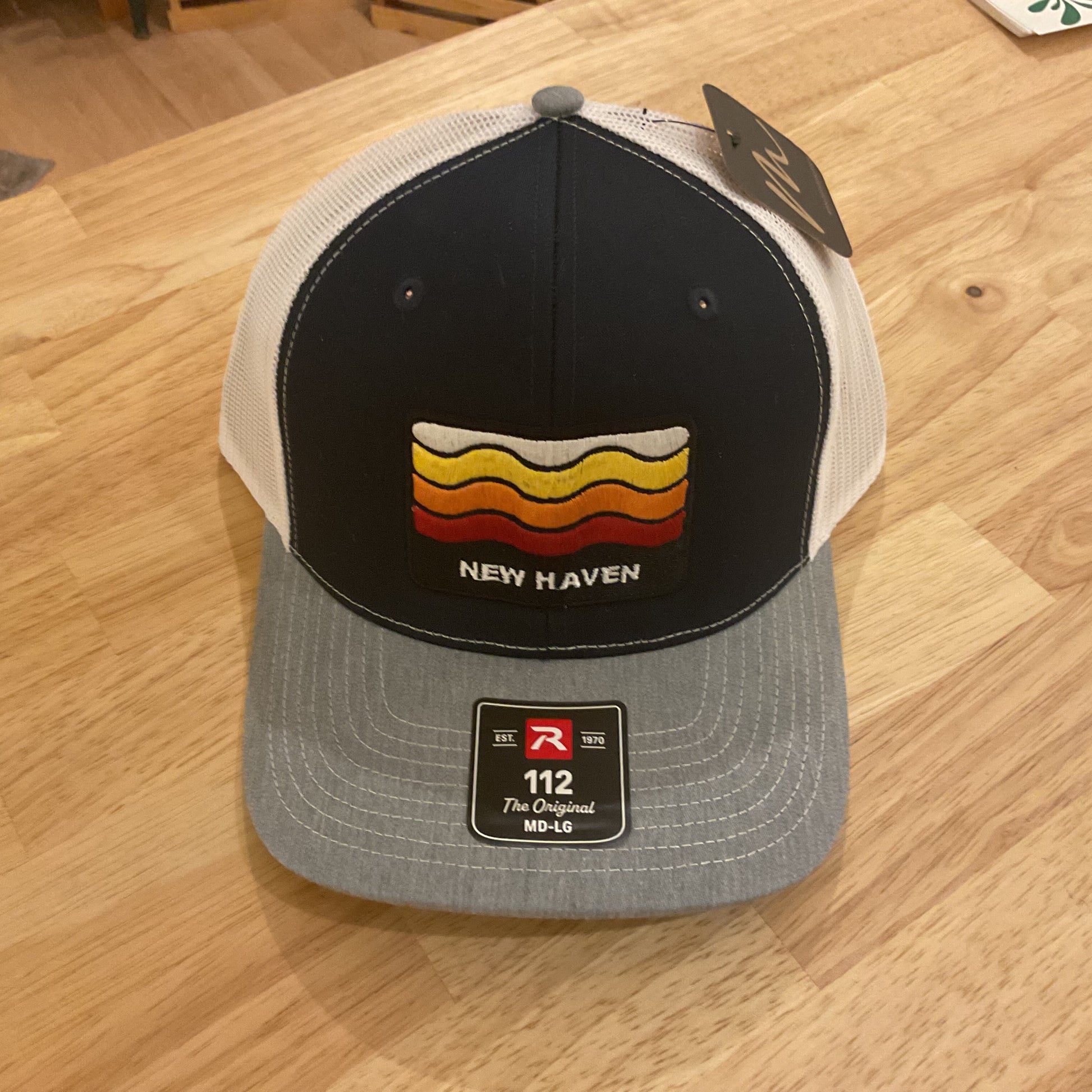 A New Haven trucker hat with a black front panel, white mesh back, and gray brim, resting on a light wood table. The front panel features a retro wavy stripe design in yellow, orange, and red with the text 'NEW HAVEN' underneath.