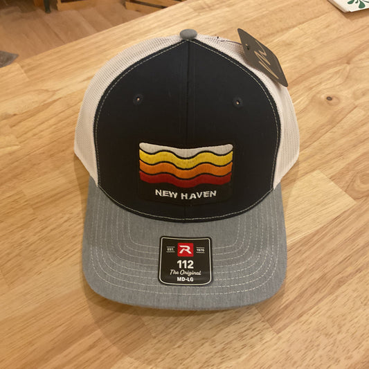 A New Haven trucker hat with a black front panel, white mesh back, and gray brim, resting on a light wood table. The front panel features a retro wavy stripe design in yellow, orange, and red with the text 'NEW HAVEN' underneath.