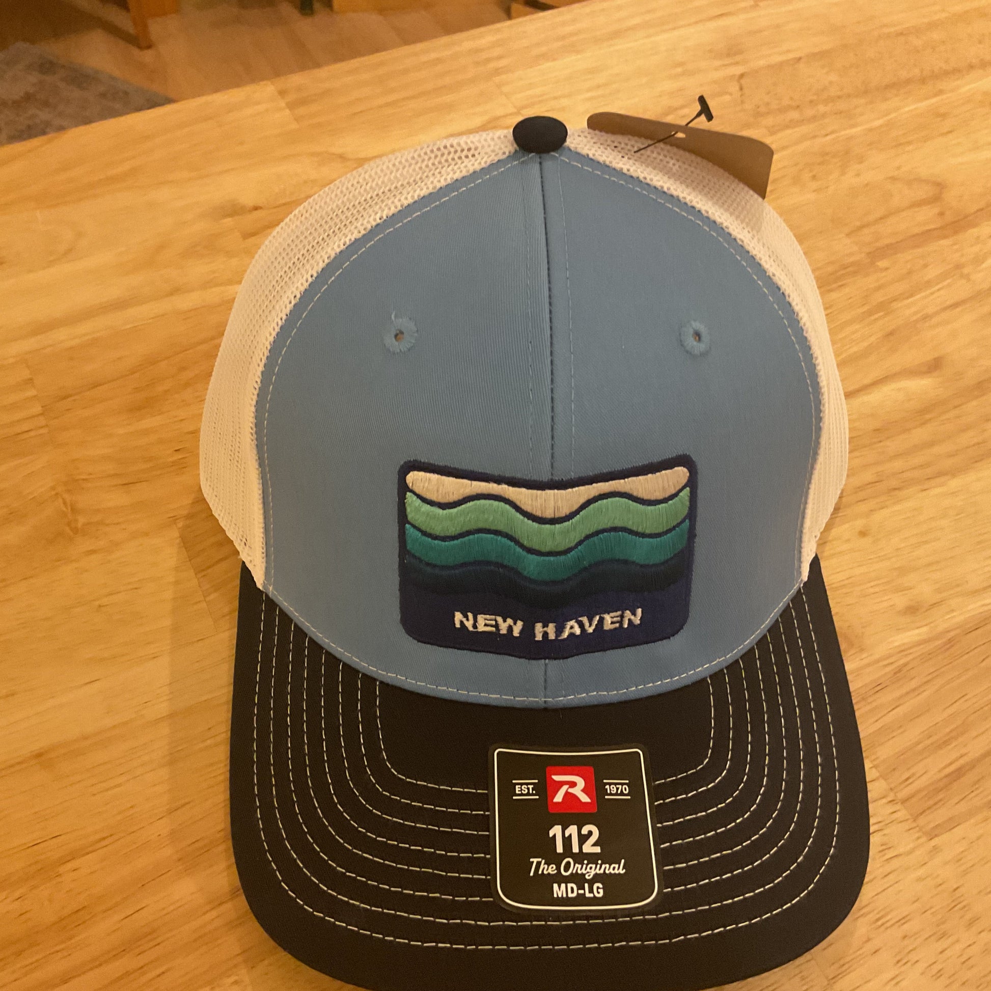 A New Haven trucker hat with a light blue front panel, white mesh back, and black visor, resting on a light wood table. The front panel features a colorful wave patch and a black size sticker.