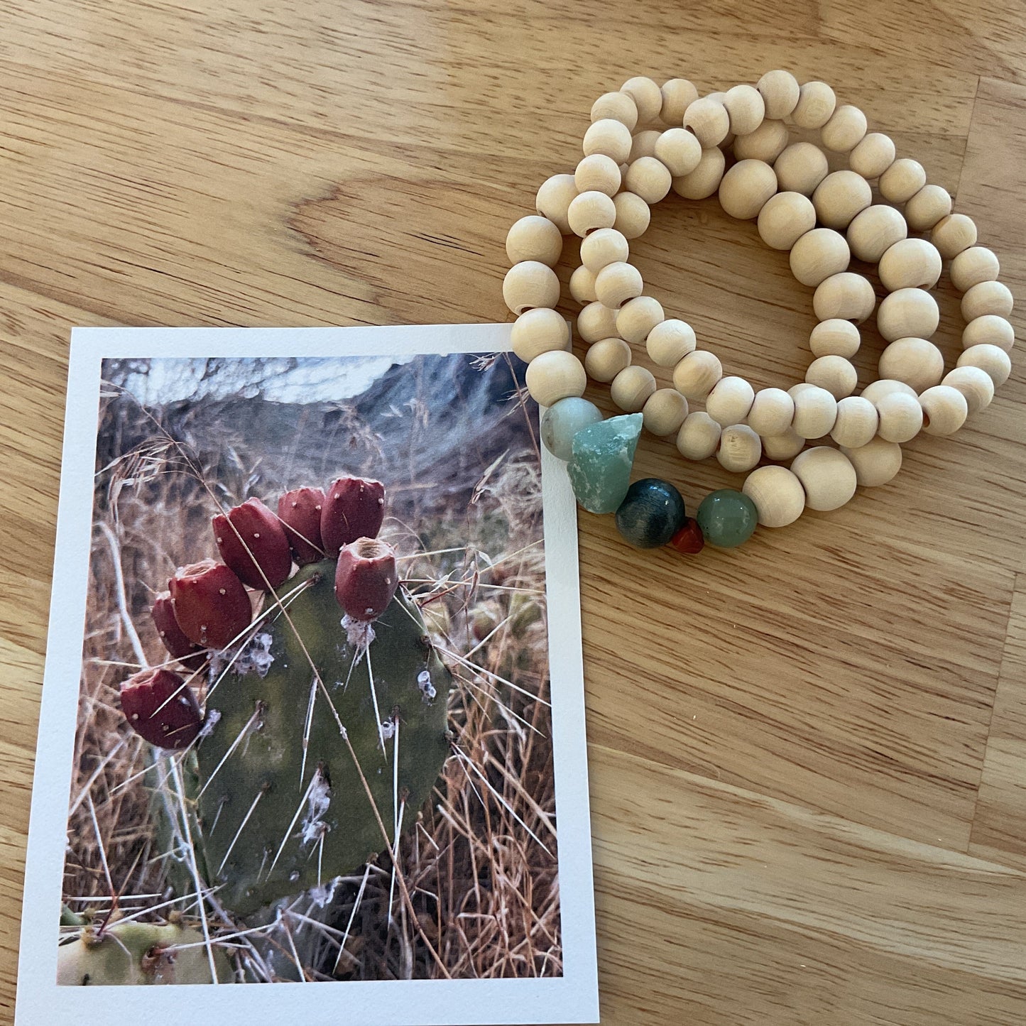 A natural wood beaded necklace with green and blue accent beads lies on a light wood tabletop next to a printed photograph of a cactus with red flowers.